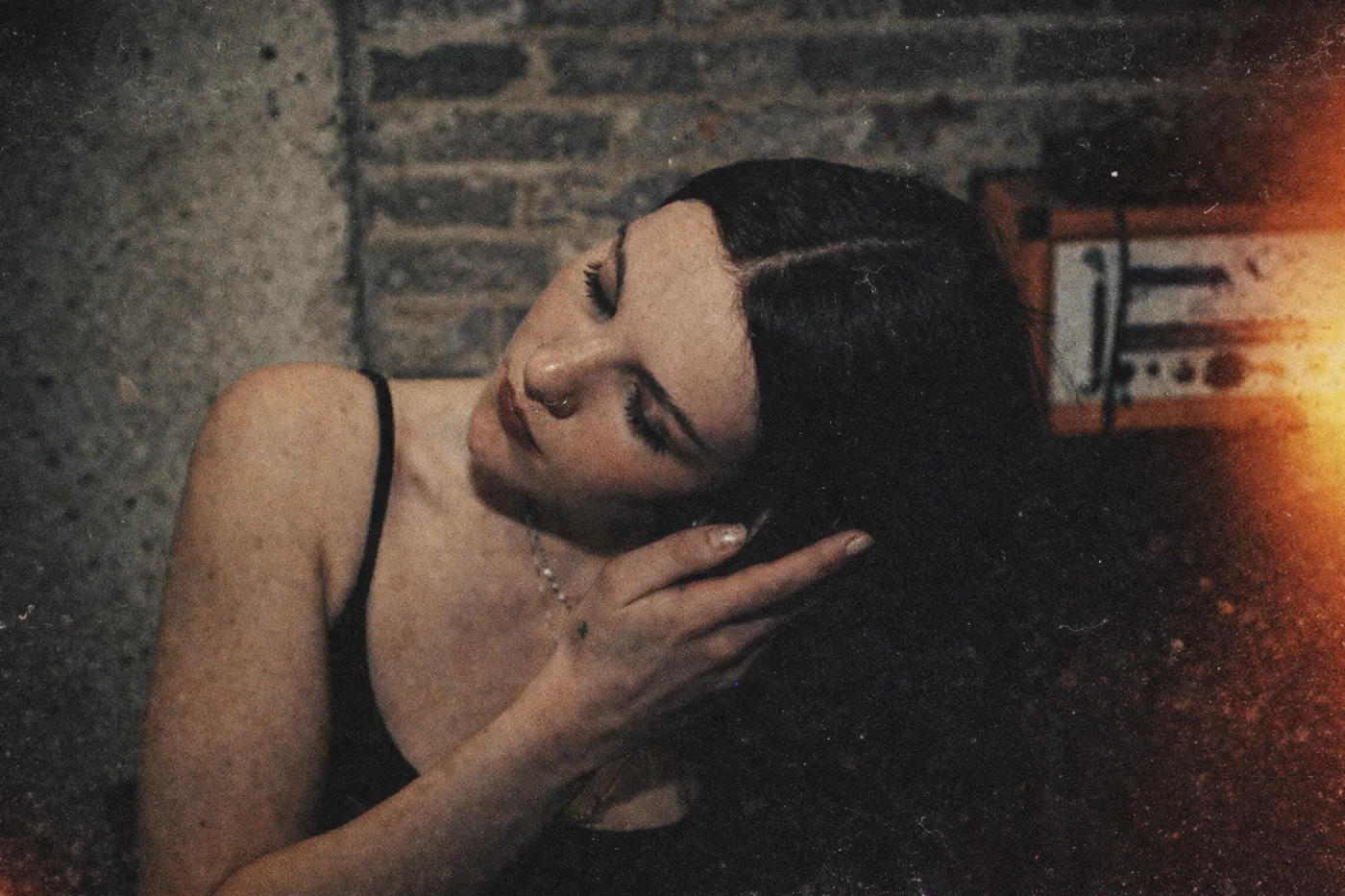 A warm, heavily grained film photograph: someone with long dark hair leaning back with their eyes closed and one hand in their hair, against a brick and concrete wall, with a hard orange light leak burning across the right of the frame.