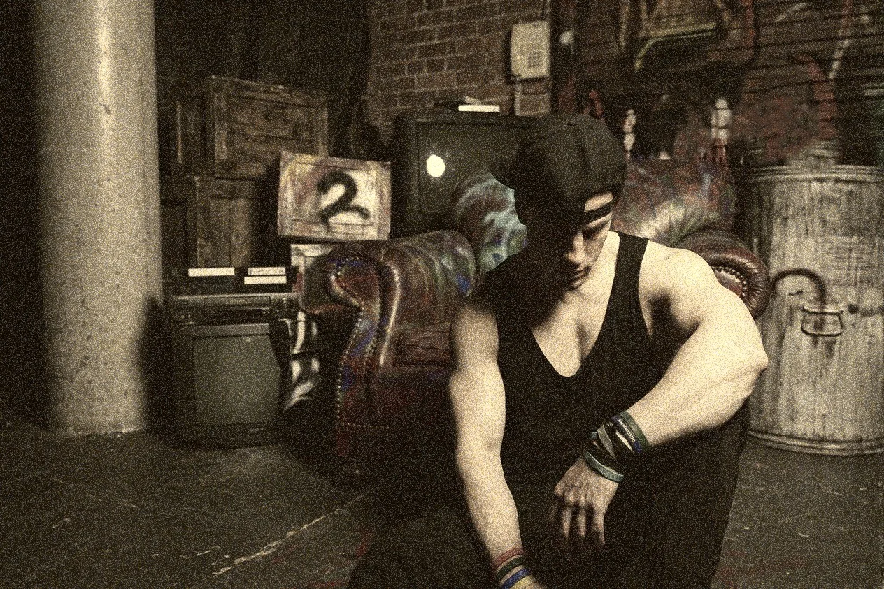 A grainy photograph in a basement-like space with a concrete pillar and brick wall: someone in a dark cap and a black tank top with wrapped wrists sitting forward on a battered leather armchair, head bowed, beside a stack of crates, an old television and a metal drum with a numbered board reading 2.