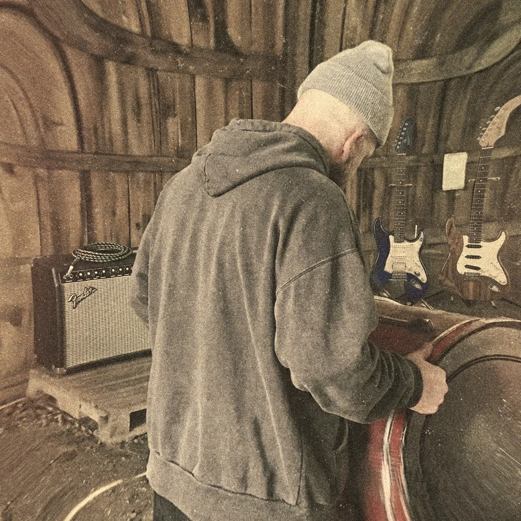 A grainy sepia photograph inside a curved wooden barn: someone in a grey hooded sweatshirt and knitted beanie seen from behind, head down and leaning over a drum, with a Fender combo amp and a coiled cable on a pallet to the left and two electric guitars hung on the boards behind.