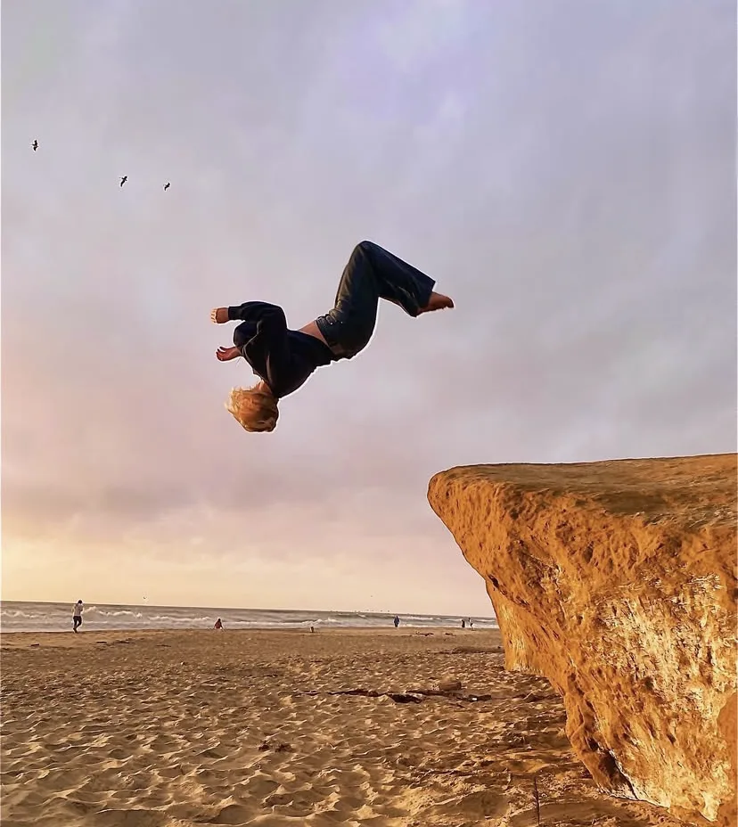 A photograph at dusk on a beach: someone in a dark jacket and jeans upside down in mid-air above the sand, having launched off a large sandstone bluff at the right of frame, with the sea, a few distant figures on the shoreline and three birds under a mauve and grey sky.