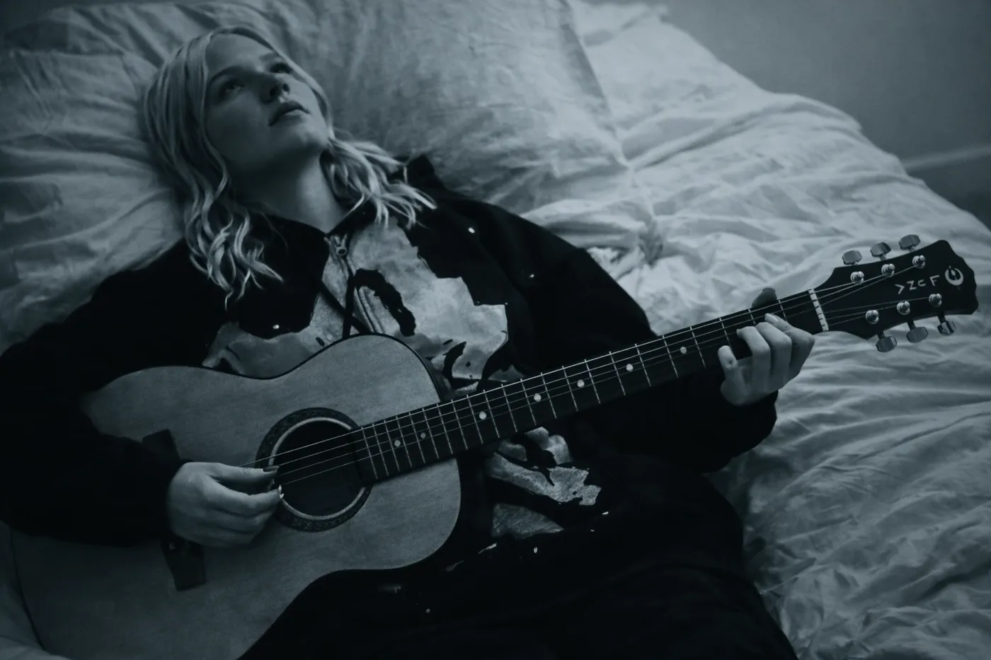 A cool-toned monochrome photograph of someone lying back across rumpled white bedding, playing an acoustic guitar and looking up out of frame, wearing a dark shirt with a distressed print.