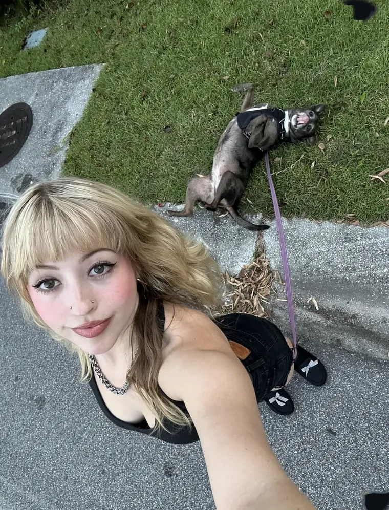 A photograph taken at arm's length looking down at a kerb: someone with a blunt fringe and a chain necklace in the foreground, and a small dog in a harness rolling on its back in the grass behind them, lead trailing across the concrete.