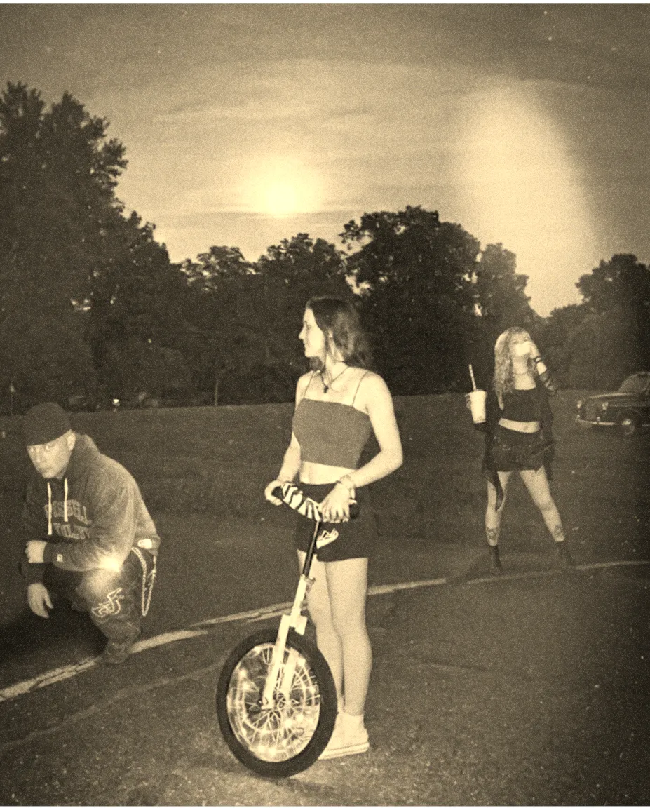 A grainy sepia photograph at dusk in an empty parking lot: one person crouching in a hooded sweatshirt and cap at the left, one standing in the centre holding a unicycle, and one at the right holding a drink cup, with a low sun behind a treeline and a parked car at the edge of frame.