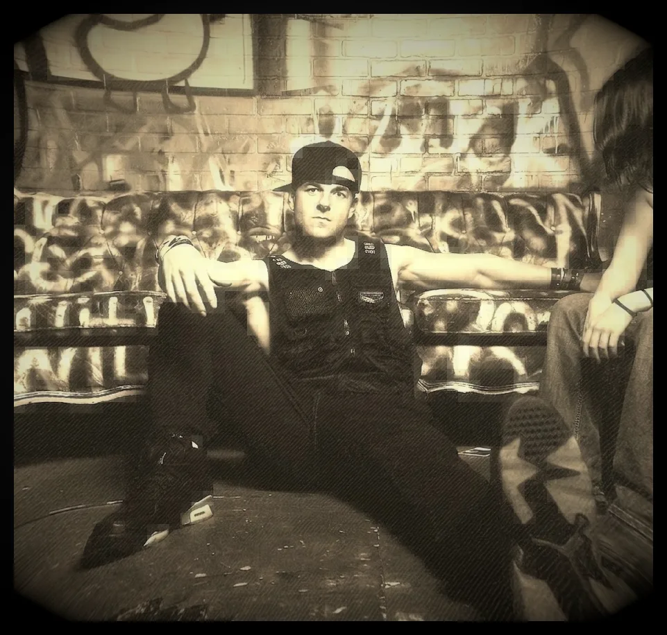 A sepia, heavily vignetted photograph in a graffiti-covered room: someone in a flat-brim cap and a dark utility vest sitting back on the floor against a spray-painted couch with one arm along the cushions, and a second person's legs and hand at the right edge of frame.