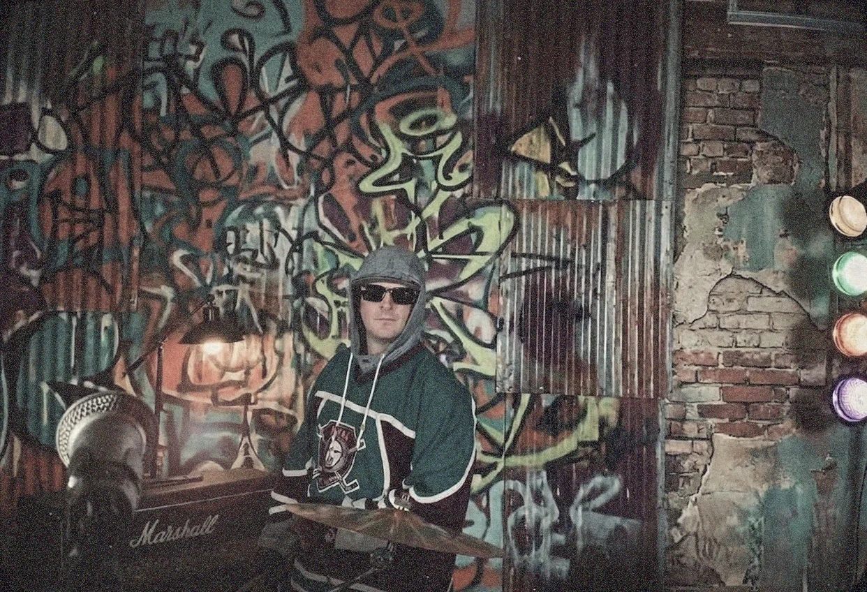 A dim colour photograph of a drummer in a hooded green hockey jersey and dark sunglasses seated behind a cymbal, in front of a wall of sprawling green and orange graffiti and corrugated metal, with a Marshall cabinet and a shaded lamp at the left and a stack of coloured traffic lights glowing at the right.
