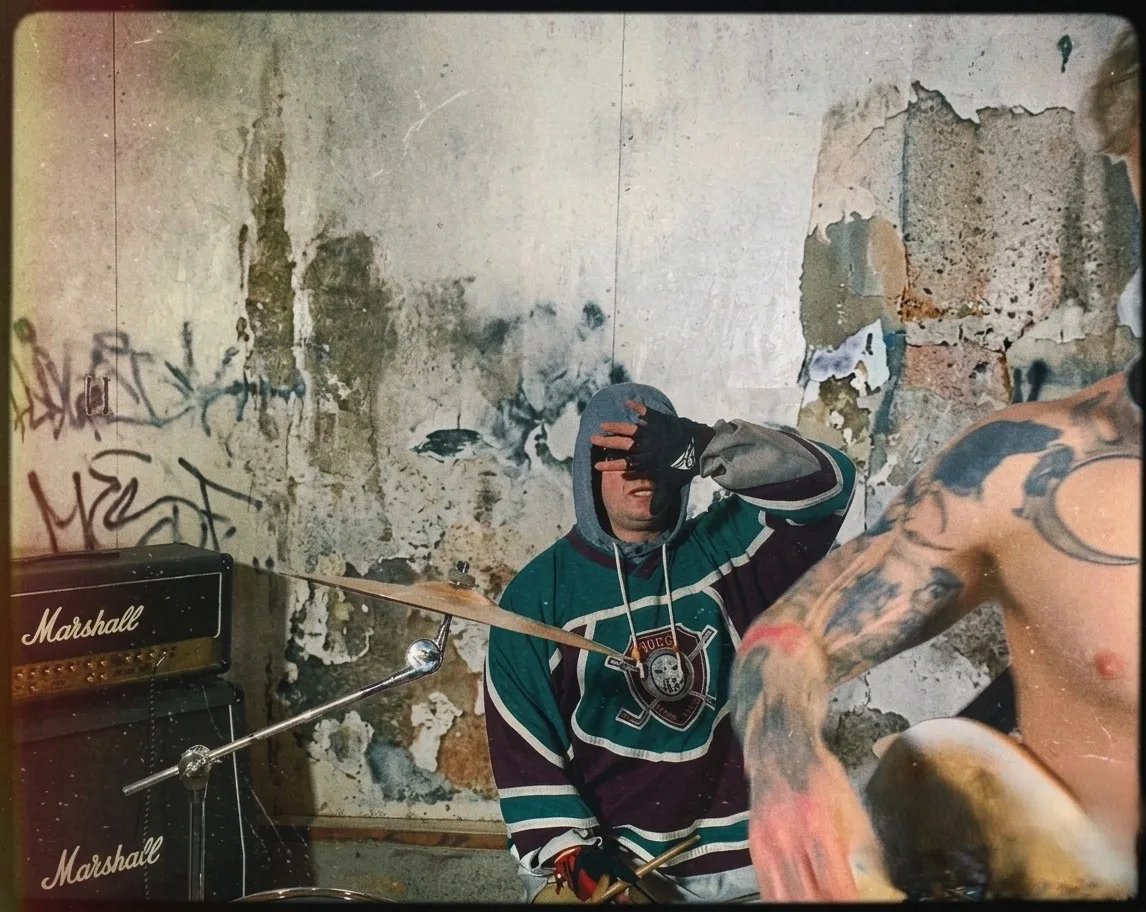 A washed-out film photograph in a derelict room with peeling plaster and graffiti: a drummer in a hooded teal and purple hockey jersey pressing a gloved hand to their forehead behind a cymbal, two stacked Marshall cabinets to the left, and a heavily tattooed shirtless figure blurred across the right of the frame.