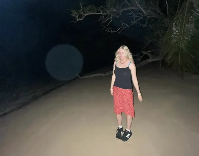 A flash photograph on a sandy path at night, palm fronds and bare branches overhead: someone standing mid-stride in a black vest top, red patterned skirt and striped trainers, with a large lens flare hanging in the dark behind them.