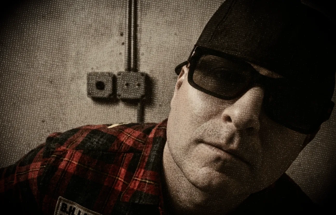 A heavily grained sepia close-up shot from below: someone in a backwards flat-brim cap and thick black sunglasses tilting their head toward the camera, wearing a red and black plaid shirt with a name patch on the chest, against a textured wall with an electrical outlet and conduit running down it.