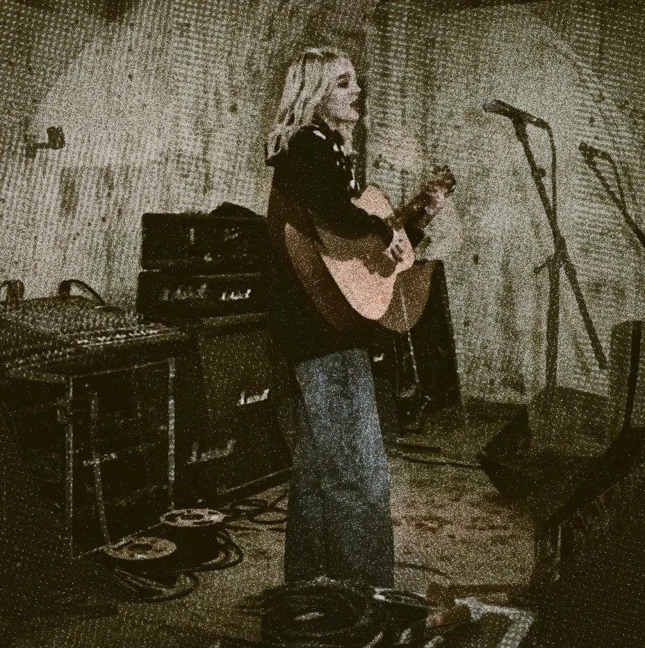 A heavily grained sepia photograph in an empty room hung with pale sheeting: someone with long light hair standing side-on playing an acoustic guitar and singing, with a mic stand, a monitor wedge, a Marshall stack and a mixing desk around them and cables coiled on the floor.