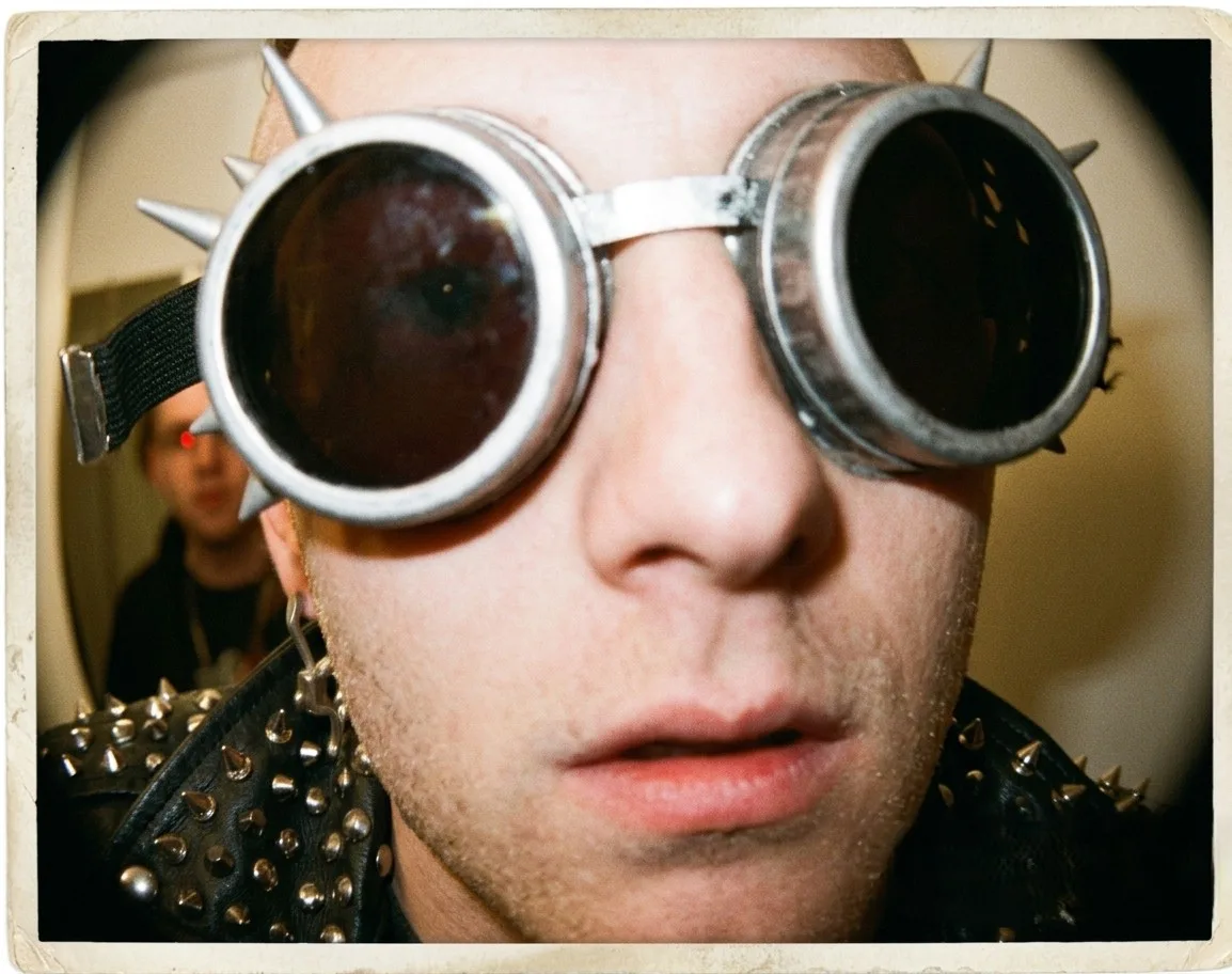 A flash-lit instant photograph with a white border, held very close: someone in spiked chrome welding goggles and a studded black leather jacket filling the frame, with a second person in the corridor behind them.