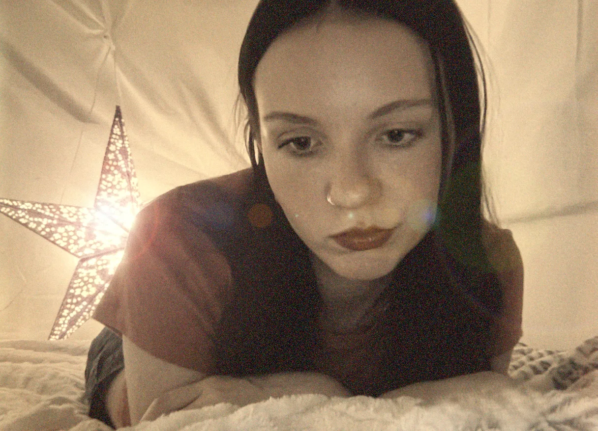 A warm, softly blown-out close-up: someone lying on their front on a bed with their chin near their folded arms, looking down out of frame, with a lit perforated star lamp flaring bright beside them against a draped pale sheet.