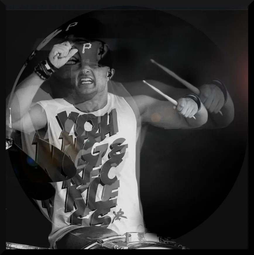 A black-and-white live photograph, ghosted with a second exposure of the same moment: a drummer in a lettered sleeveless shirt and a cap mid-strike behind a kit, both sticks raised, face screwed up with effort.