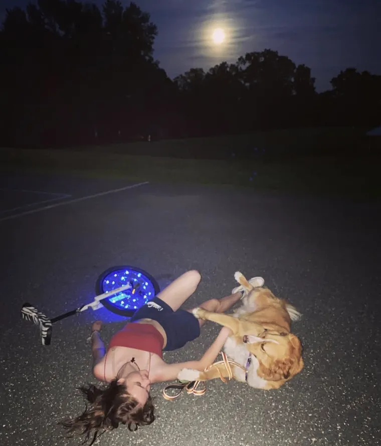 A night photograph on empty asphalt under a full moon: someone lying flat on their back beside a dog that is also lying down, with a unicycle on its side next to them, its wheel ringed with blue LEDs.